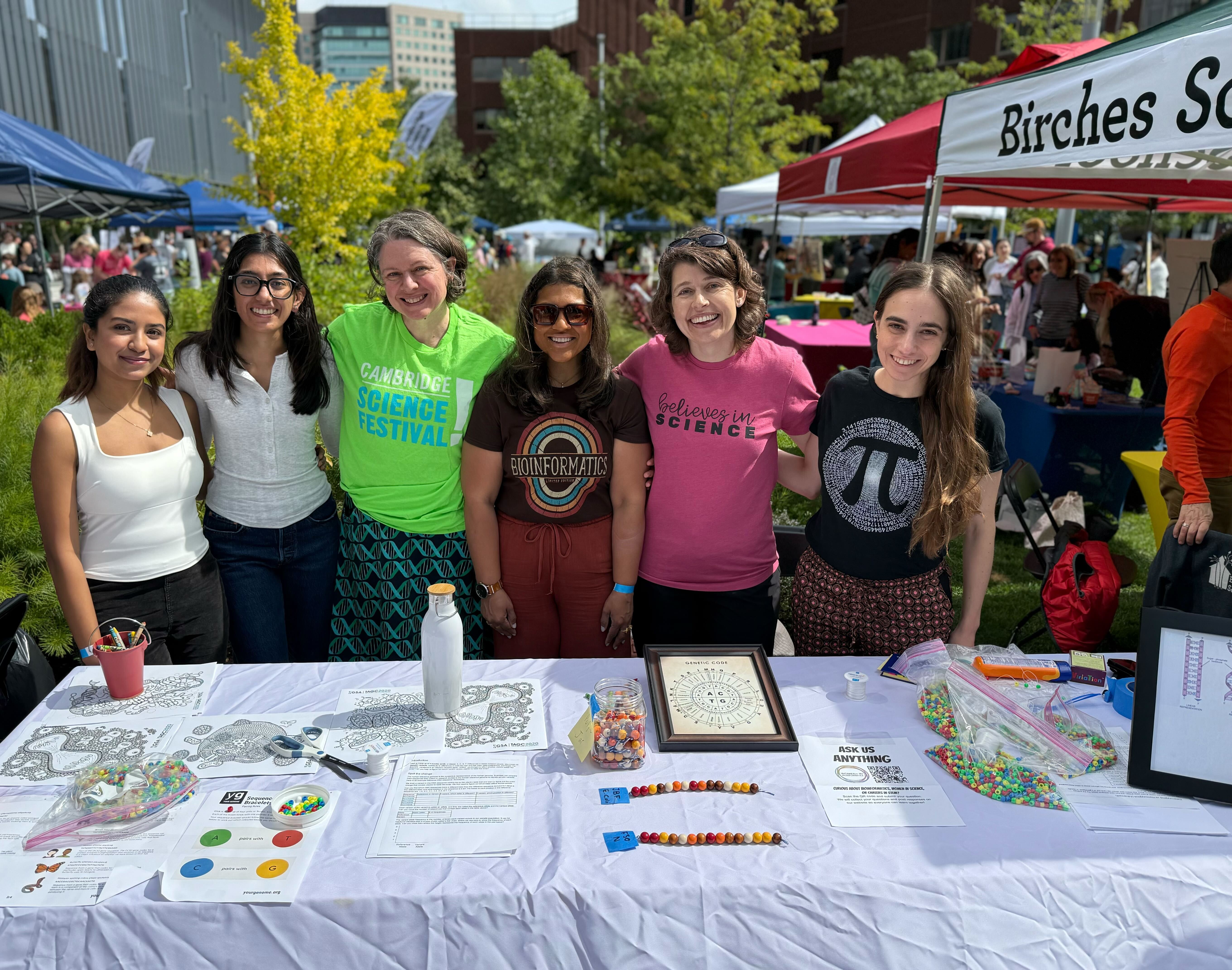 Six women at Cambridge Science Festival booth with kids' activity table. Shirts include "Cambridge Science Festival," "Bioinformatics" with rainbow, "Believes in Science," and pi symbol.